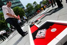 As the sun beat down on Regent’s Place last Thursday and Friday, the plaza was transformed into a golf fest for the masses. For two days only anyone could come and play for free on our portable 9-hole ‘Signature Golf Course’, soak in the rays on the temporary deck chairs and enjoy the Open Championship Golf on the jumbo screen. The activity attracted all kinds of visitors, but mostly it was staff from the surrounding office buildings and people passing through on their lunch break. There was also a scattering of tourists, day trippers and commuters. But everyone had one thing in common – a smile on their face! Our client dressed the area with rainbow-coloured beach huts, banners and bright orange statues. And with the BT Tower looming in the background it really was a sight to see. Click on the ‘enlarge’ button to the right for a better look at the pictures. The crazy golf course included two special holes. One was customised for our client to include their logo and brand colours. The other was our brand new red and black ‘Casino’-themed hole on which players were tempted to gamble on going for a hole-in-one. This was my first event since I began working for UrbanCrazy and it was hugely exciting for me to be involved in. However, there was a slightly embarrassing moment, when, while attempting an ace our ‘DreamGolf’ track, the ball rebounded right back to my feet. Oh how we laughed! But I had the last laugh, when on my next attempt I got it straight in. We were delighted to hear positive feedback from our client, Regent’s Place. They said “UrbanCrazy delivered two days of crazy fun on the plaza. Alongside The Open Championship, the crazy golf was a huge success”. To make your next event a huge success, look no further than UrbanCrazy’s colourful and quirky ‘Signature Course’. I guarantee it will make you smile!